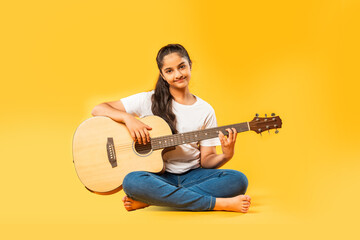 Full body portrait of Indian asian small girl while playing guitar against yellow background