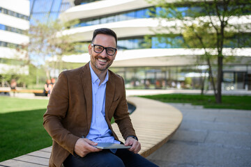 Portrait of happy male employee with laptop sitting on bench against modern office buildings in city