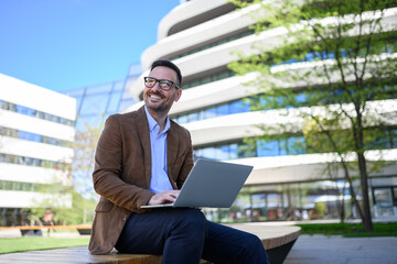Low angle view of smiling man working over laptop and looking away while sitting on bench in city
