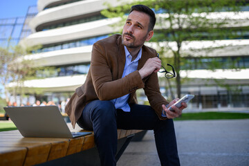 Thoughtful young businessman with cellphone and laptop looking away while sitting on bench in city