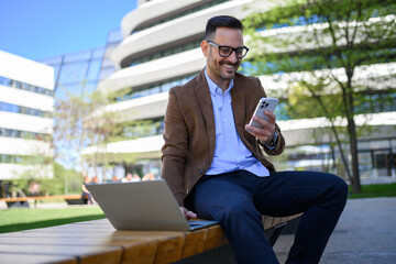Handsome entrepreneur messaging over cellphone and using laptop while sitting on bench in city