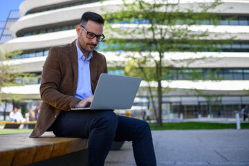 Focused businessman analyzing financial report over laptop while sitting on bench in modern city