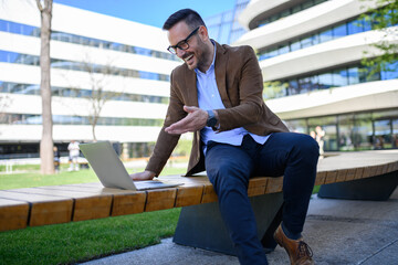 Positive entrepreneur discussing over video call on laptop while sitting on bench against buildings