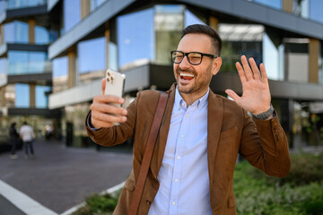 Cheerful young man in glasses waving hand over video call on cellphone while standing in modern city