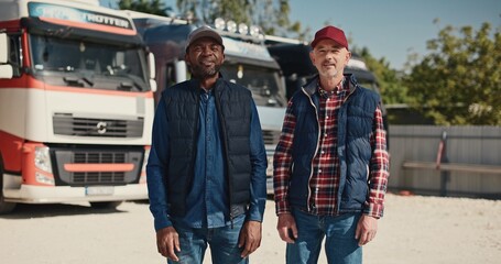 Obraz premium Two mixed-race coworkers standing before large trucks outside. Mature drivers in hats and jackets positively looking into camera. Getting ready to traveling. Working together in delivery company.