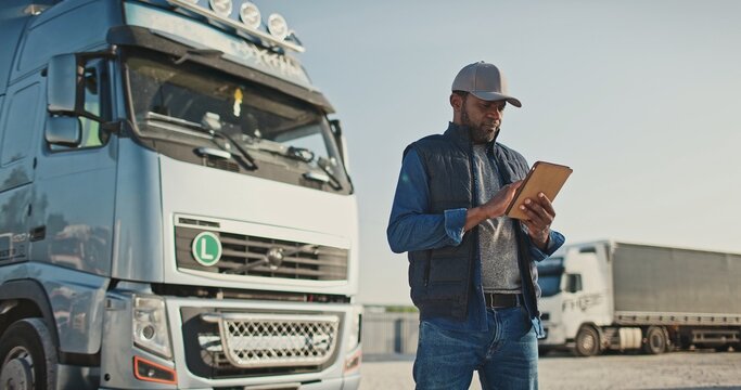 African American professional truck driver setting up navigation for destination. Checking his route on tablet computer and standing by long vehicle. Transportation service.