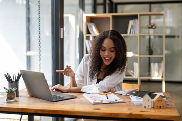 A woman is sitting at a desk with a laptop and pointing at something on the screen. She is smiling and she is happy. The scene suggests a positive and productive atmosphere
