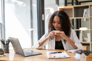 A woman is sitting at a desk with a laptop and a cup of coffee. She is smiling and she is enjoying her coffee