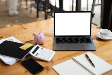 A laptop is on a table with a pink piggy bank, a calculator, a pen, and a cup. The table is...