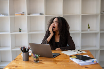 A young woman yawns while using a laptop at her desk. being tired during a busy work day
