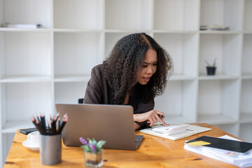 A woman is sitting at a desk with a laptop and a notebook. She is looking at the notebook and pointing at something. The scene suggests that she is working on a project or trying to solve a problem