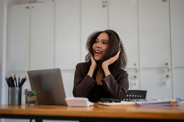 A woman with curly hair is sitting at a desk with a laptop and smiling. She is happy and excited about something
