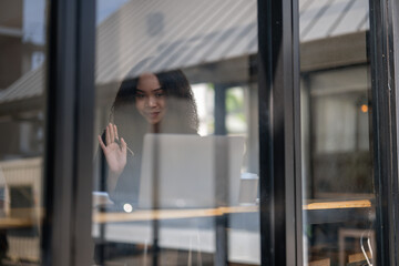 A woman is looking out a window at a laptop. She is waving to someone on the other side of the...