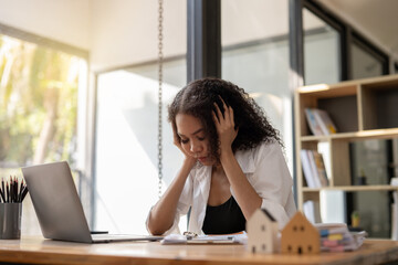 A woman is sitting at a desk with a laptop and a stack of papers. She is looking at the laptop and she is in a state of distress