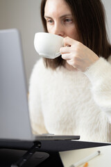 Focused woman drinking coffee in front of laptop screen. Using laptop stand for comfortable working. Busy businesswoman, home office, online meeting, shopping online, overtime working