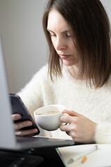 Woman looking serious at cell phone, holding cup of coffee and sitting in front of laptop screen. Blogging, surfing social media, home office, online meeting, stock trading, analysis data