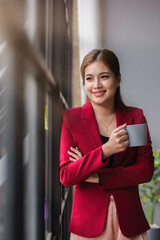 Confident smiling business woman stands holding a coffee mug and looks out the window.