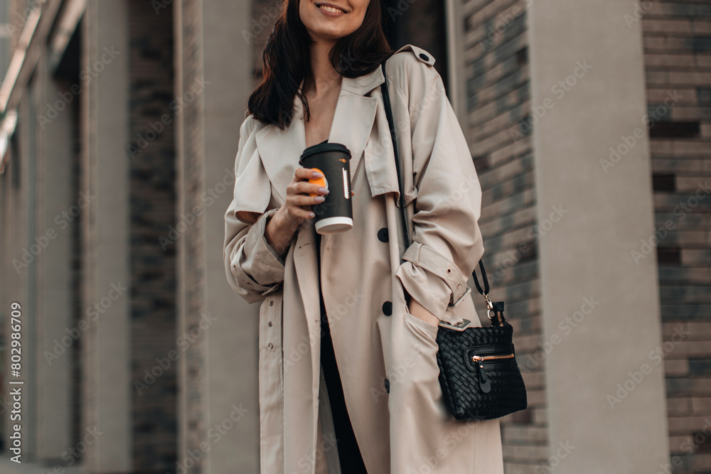 Wall mural Fashion details of beige trench coat, woman walking and drinking coffee against the background of the city. Street style cloth