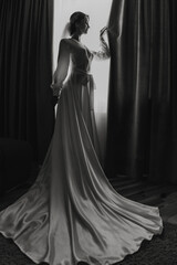 A woman in a wedding dress stands in front of a curtain. The dress is long and flowing, and the...
