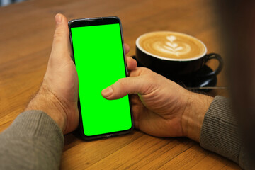 Young man sitting at cafe holding smartphone green mock-up screen in hand. Male person using chroma...