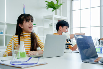 Students' learning in science. Focused girl in yellow-striped top engages with science experiment, observing green liquid in flask, laptop open on table in classroom.