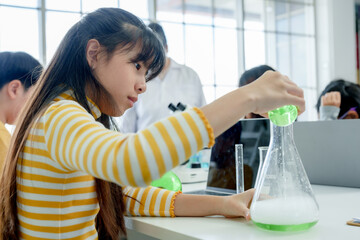 Students' learning in science classroom. Focused girl in yellow-striped shirt pours green liquid from test tube into flask, with microscope and classmates in background, in lab classroom. - Powered by Adobe