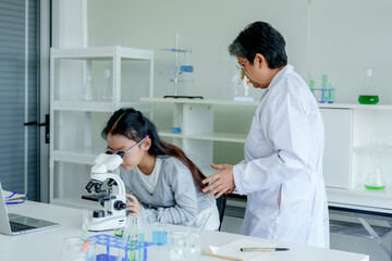 Obraz premium Students' learning in science classroom. Asian educator assists young girl in gray hoodie, learning to use microscope in science classroom with laptop and lab equipment on desk.