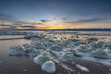 Sea foam washes up on the beach of Den Helder near a pier during a colorful sunset. Foam on...