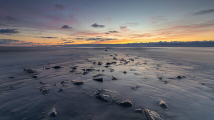 A vast view of the North Sea, where the pier slowly emerges as the sea recedes during a colorful sunset. The peak of the tide has passed, revealing the pier gradually. Colourful clouds in the sky.