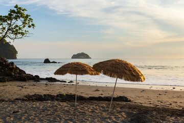 Two straw parasols planted on deserted beach seen at dusk, with small island in the Pacific Ocean...