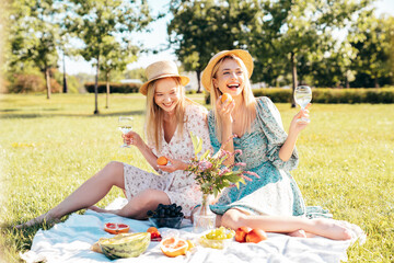 Two young beautiful smiling hipster female in summer sundress and hats. Carefree women making picnic outside. Positive models sitting on plaid on grass, drinking champagne,  eating fruits