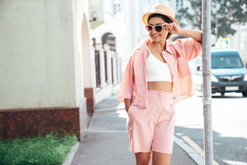 Fototapeta premium Young beautiful smiling hipster woman in trendy summer pink costume clothes. Carefree female posing in the street at sunny day. Positive model outdoors at sunset. Cheerful and happy in hat, sunglasses