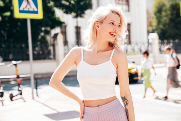 Young beautiful smiling blond hipster woman in trendy summer clothes. Carefree female posing in the street at sunny day. Positive model outdoors at sunset. Cheerful and happy