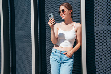 Young beautiful smiling hipster woman in trendy summer casual clothes. Carefree woman posing in the street. Positive model holds mobile phone, looks at cellphone screen, uses smartphone apps