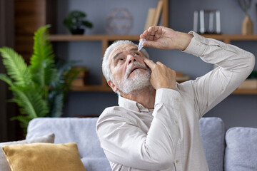 An elderly retired man is at home, sitting on the sofa and instilling medicine in his eye for inflammation and fatigue. The photo is close