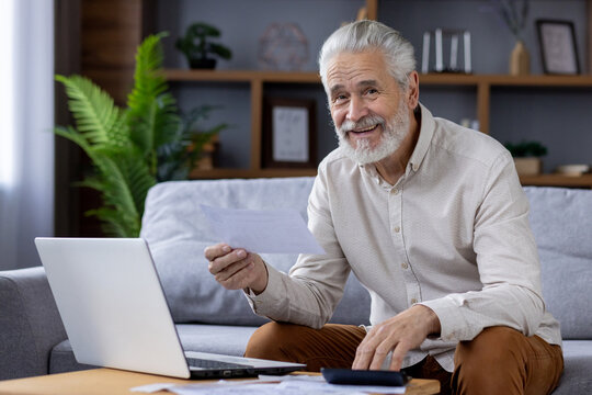 Portrait of smiling senior gray-haired man sitting on sofa at home and checking bills and documents, using laptop and calculator, looking at camera