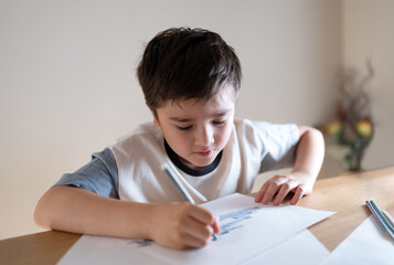 Kid siting on table doing homework,Child boy holding grey pen clouring and drawing on white paper,...