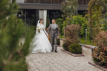 A bride and groom are walking down a path in a garden. The bride is wearing a white dress and the...
