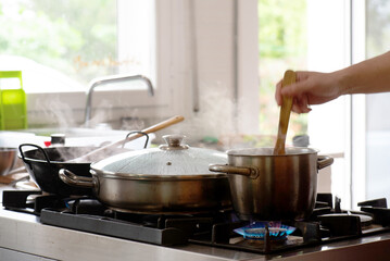hand stirring a pot, kitchen full of pots cooking and cooking with gas, in the background a window with natural light and vegetation.