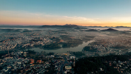 Aerial view of Dalat City at sunrise, featuring the tranquil Xuan Huong Lake amidst urban structures, surrounded by lush greenery and misty hills under a golden sky.