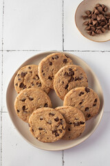 Plate of chocolate chip cookies with extra chocolate chips on side, set on a tiled background