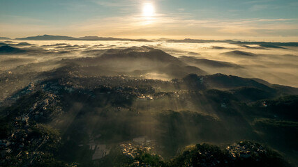 Aerial view of Da Lat at sunrise, showcasing mist-covered buildings amidst lush mountains, under a golden sky, highlighting the city’s architectural and natural beauty.