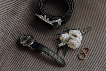 A watch, a belt, and a boutonniere are displayed on a gray surface. The watch is black and has a silver band - obrazy, fototapety, plakaty