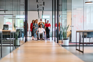 A diverse group of young business people walking a corridor in the glass-enclosed office of a modern startup, including a person in a wheelchair and a woman wearing a hijab