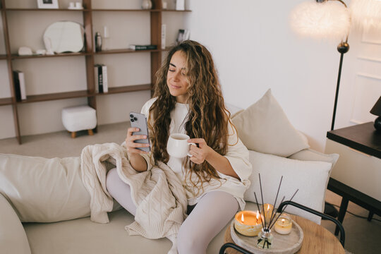 Young woman using her smartphone and drinking tea, relaxing in a cozy living room.