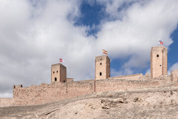 An ancient stone castle atop a hill, with three towers and flags, under a partly cloudy sky