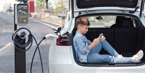 Little boy sitting on car trunk, using smartphone while recharging eco-friendly car from EV...