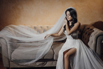 A woman in a white wedding dress sits on a couch with a veil draped over her head. The couch is...