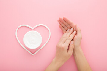 Young adult woman hands and white heart shape with cream jar on light pink table background. Pastel...