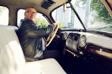 elderly man dressed in a black jacket on a city street driving a vintage car